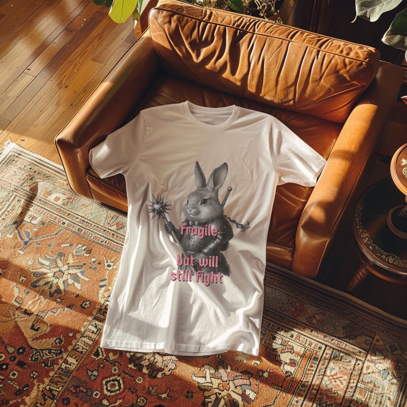 K&ouml;nnte beinhalten: Wei&szlig;es T-Shirt mit einer Illustration eines Kaninchens in R&uuml;stung, das eine Waffe und ein Schwert h&auml;lt. Darunter steht in Rosa "fragile but will still fight". Das Shirt liegt auf einem gemusterten Teppich und einem Ledersessel.