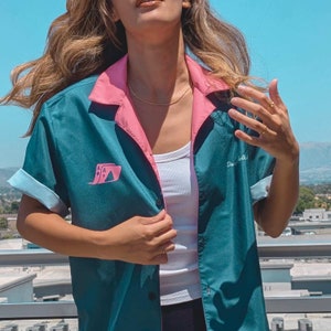 May include: A teal blue bowling shirt with pink trim and a playing card graphic. The shirt has a white button-up front and a white tank top underneath.