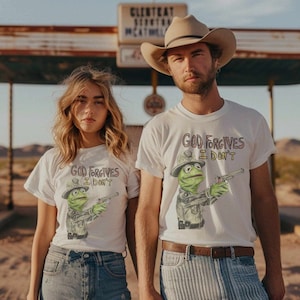 May include: Two people wearing white t-shirts with a green Muppet character holding a gun and the text "God Forgives I Don't". The person on the left is wearing blue jeans and the person on the right is wearing a brown cowboy hat and striped blue jeans.
