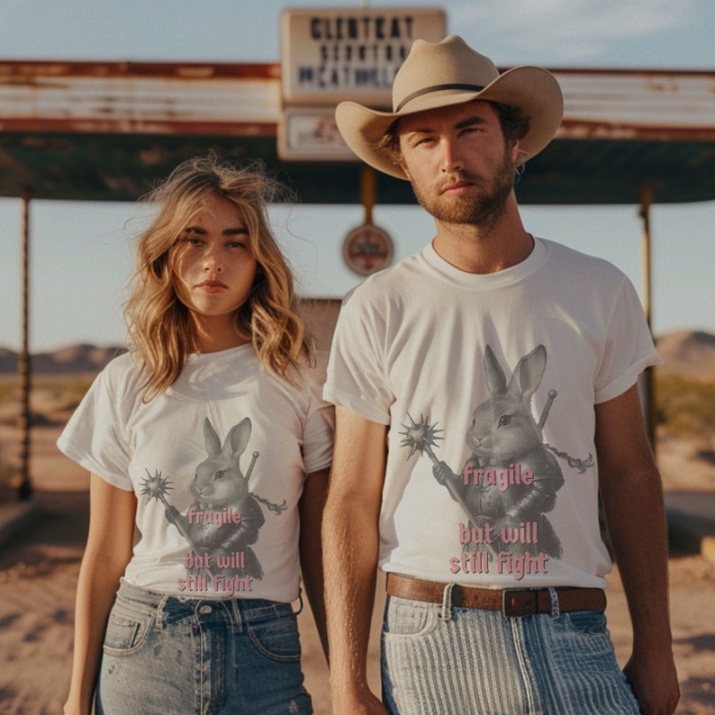 K&ouml;nnte beinhalten: Wei&szlig;e T-Shirts mit einem Kaninchen-Krieger-Motiv und dem Text "fragile but will still fight". Das Kaninchen h&auml;lt eine Keule und ein Schwert. Die Models tragen blaue Jeans und einen Cowboyhut.