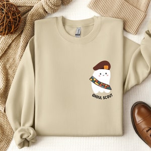 May include: A tan sweatshirt with a cartoon ghost wearing a scout hat and sash. The text "Ghoul Scout" is printed below the ghost.
