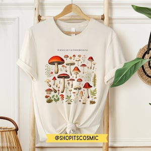 May include: A white t-shirt with a vintage illustration of various mushrooms. The illustration is in shades of red, brown, and green. The text "Science of the North American" is printed above the illustration. The text "@shopitscosmic" is printed below the illustration.