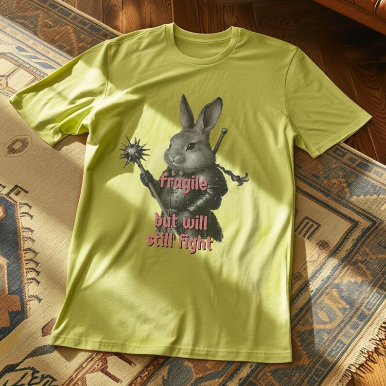 K&ouml;nnte beinhalten: Hellgr&uuml;nes T-Shirt mit einer Illustration eines Kaninchenkriegers in R&uuml;stung, der eine Keule und ein Schwert h&auml;lt. Der Text "fragile but will still fight" ist in Rosa gedruckt. Das T-Shirt ist aus weichem Material.