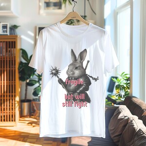 K&ouml;nnte beinhalten: Wei&szlig;es T-Shirt mit einem Design eines Kaninchens in R&uuml;stung, das eine Keule und ein Schwert h&auml;lt. Der rosa Text lautet "fragile but will still fight". Das T-Shirt h&auml;ngt an einem Holzb&uuml;gel.
