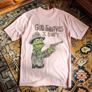 May include: Pink t-shirt with a graphic of a green Muppet character wearing a police hat and holding a gun. The text "GOD FORGIVES I DON'T" is printed on the shirt.
