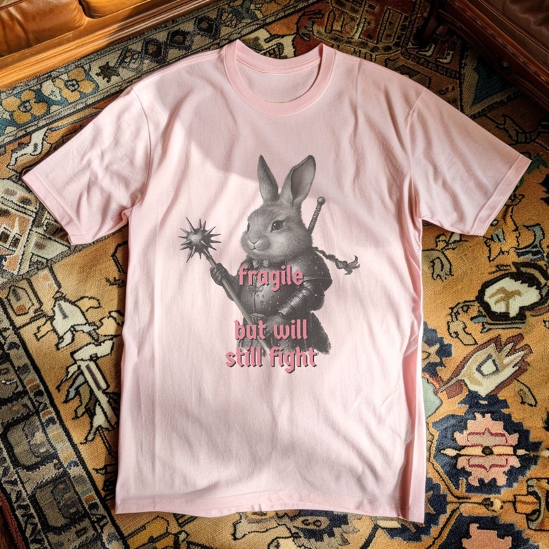 K&ouml;nnte beinhalten: Hellrosa T-Shirt mit detaillierter Illustration eines Kaninchens in R&uuml;stung, das eine Keule und ein Schwert h&auml;lt. Der Text "fragile but will still fight" ist unter dem Kaninchen in Rosa gedruckt. Das Shirt ist aus weichem Material.
