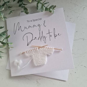 May include: A white card with the words "To a Special Mummy & Daddy to be" in cursive script. A small, white knitted sweater is attached, complete with knitting needles and a ball of yarn, perfect for a new parent.