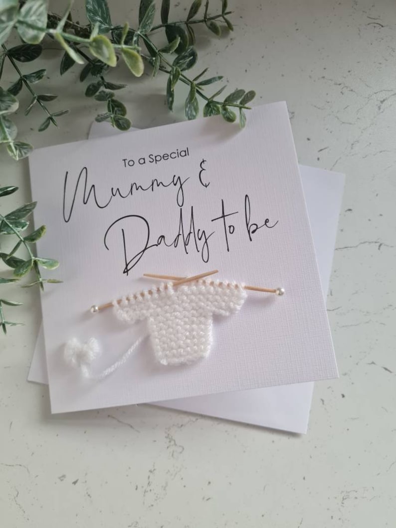 May include: A white greeting card with the text "To a Special Mummy & Daddy to be" in elegant script. A miniature white knitted sweater with knitting needles and a ball of yarn is attached to the card, suggesting a baby theme.