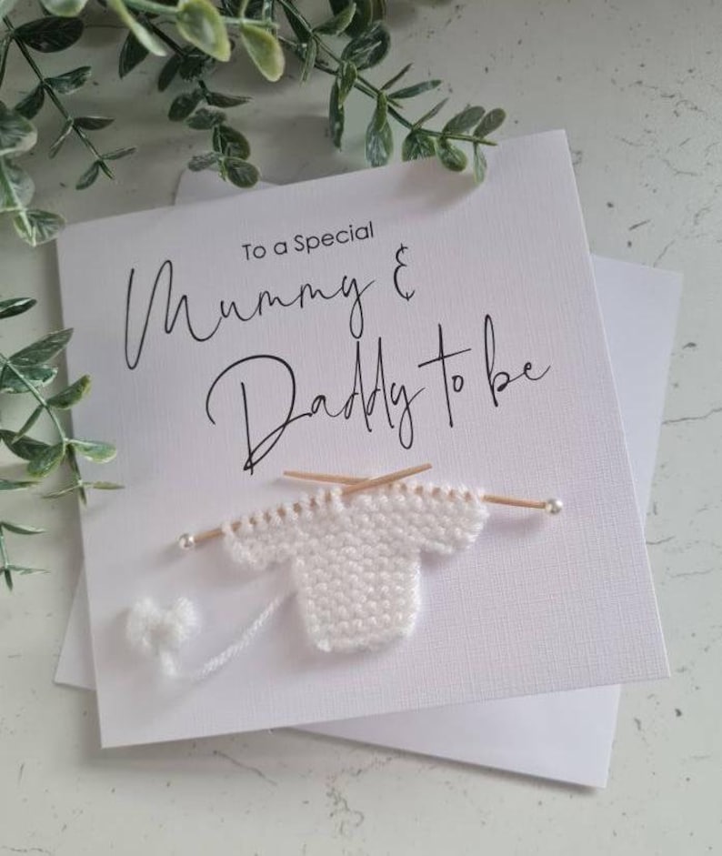 May include: A white card with black text that reads "To a Special Mummy & Daddy to be". A small white knitted sweater is attached to the card with two wooden knitting needles.