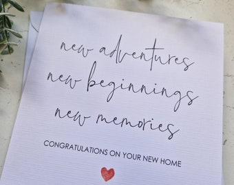 May include: A white card with black calligraphy that reads "new adventures new beginnings new memories" and "Congratulations on your new home". A red heart is in the bottom right corner.
