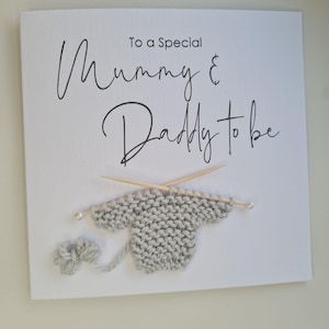 May include: A white card with the text "To a Special Mummy & Daddy to be" in black script. A gray knitted sweater with wooden knitting needles and a ball of yarn are attached to the card.