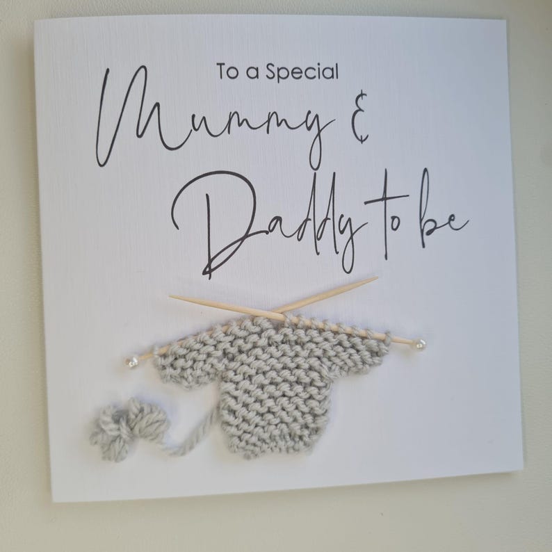 May include: A white card with the text "To a Special Mummy & Daddy to be" in black script. A gray knitted sweater with wooden knitting needles and a ball of yarn are attached to the card.