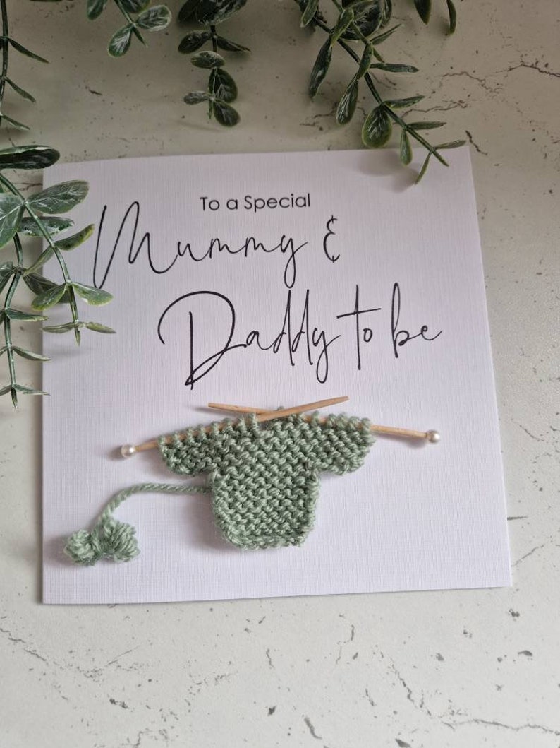 May include: A white card with the text "To a Special Mummy & Daddy to be" in black script. A small green knitted sweater is attached to the card with two wooden knitting needles.