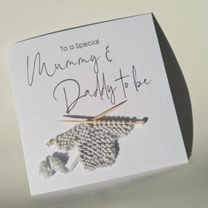 May include: A white card with the text "To a Special Mummy & Daddy to be" in elegant script. The card features a small, gray knitted sweater with knitting needles and a ball of yarn, suggesting a baby theme.