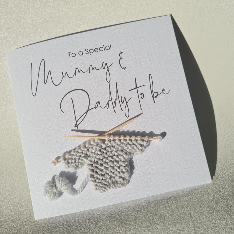 May include: A white card with the text "To a Special Mummy & Daddy to be" in elegant script. The card features a small, gray knitted sweater with knitting needles and a ball of yarn, suggesting a baby theme.