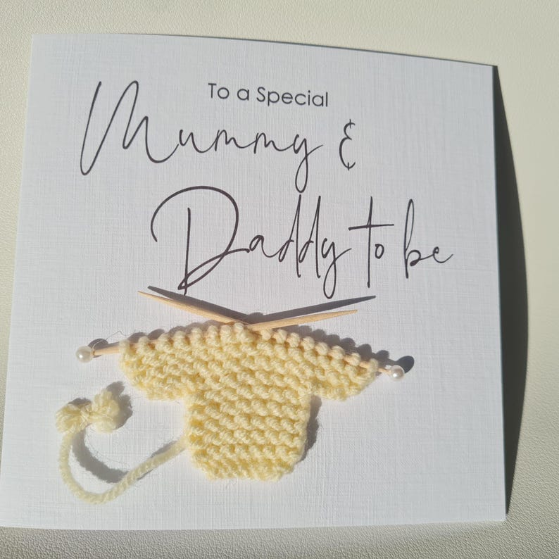 May include: A white card with the text "To a Special Mummy & Daddy to be" in black script. A small, knitted yellow sweater is attached, with knitting needles and a pearl accent.