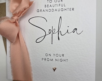 May include: A white card with the text "TO OUR BEAUTIFUL GRANDDAUGHTER Sophia ON YOUR PROM NIGHT" is adorned with a peach-colored ribbon and bow. A small gold heart is at the bottom. The card is on a brown envelope.