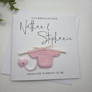 May include: A white greeting card with the names "Nathan & Stephanie" in elegant script. The card features a small, pink knitted sweater with knitting needles and the text "AMAZING PARENTS TO BE".