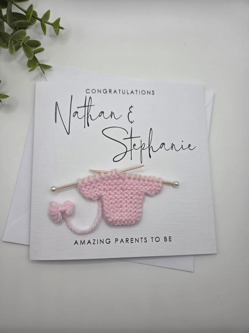 May include: A white greeting card with the names "Nathan & Stephanie" in elegant script. The card features a small, pink knitted sweater with knitting needles and the text "AMAZING PARENTS TO BE".