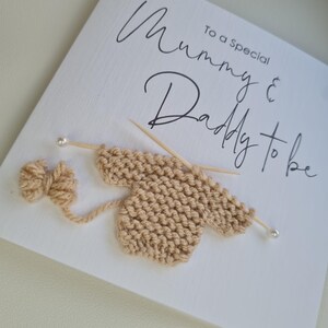May include: A white card with the text "To a Special Mummy & Daddy to be" in black script. A beige knitted baby sweater is depicted with knitting needles and a yarn bow, suggesting a baby shower or pregnancy announcement.