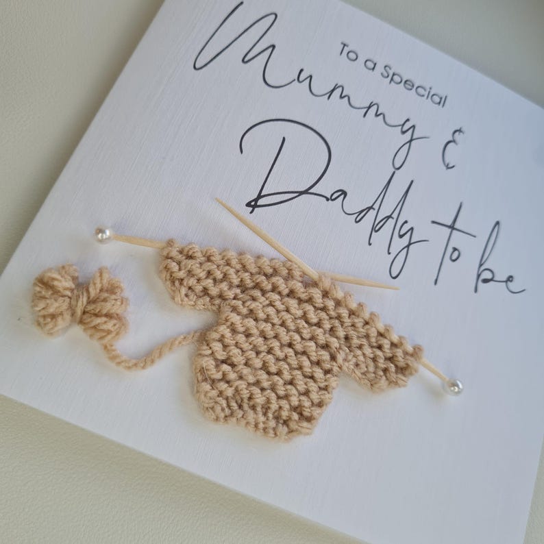 May include: A white card with the text "To a Special Mummy & Daddy to be" in black script. A beige knitted baby sweater is depicted with knitting needles and a yarn bow, suggesting a baby shower or pregnancy announcement.