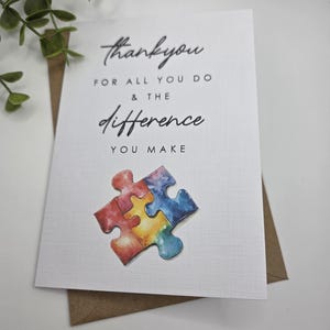 May include: A white greeting card with the words "thank you for all you do & the difference you make" in black script. A watercolor puzzle piece in red, yellow, blue, and purple is at the bottom. The card is on a brown envelope.