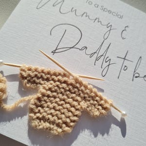 May include: A handmade greeting card with the words "To a Special Mummy & Daddy to be" in elegant script. The card features a small, knitted beige baby garment with knitting needles and pearl-tipped pins, suggesting a baby shower or pregnancy announcement.