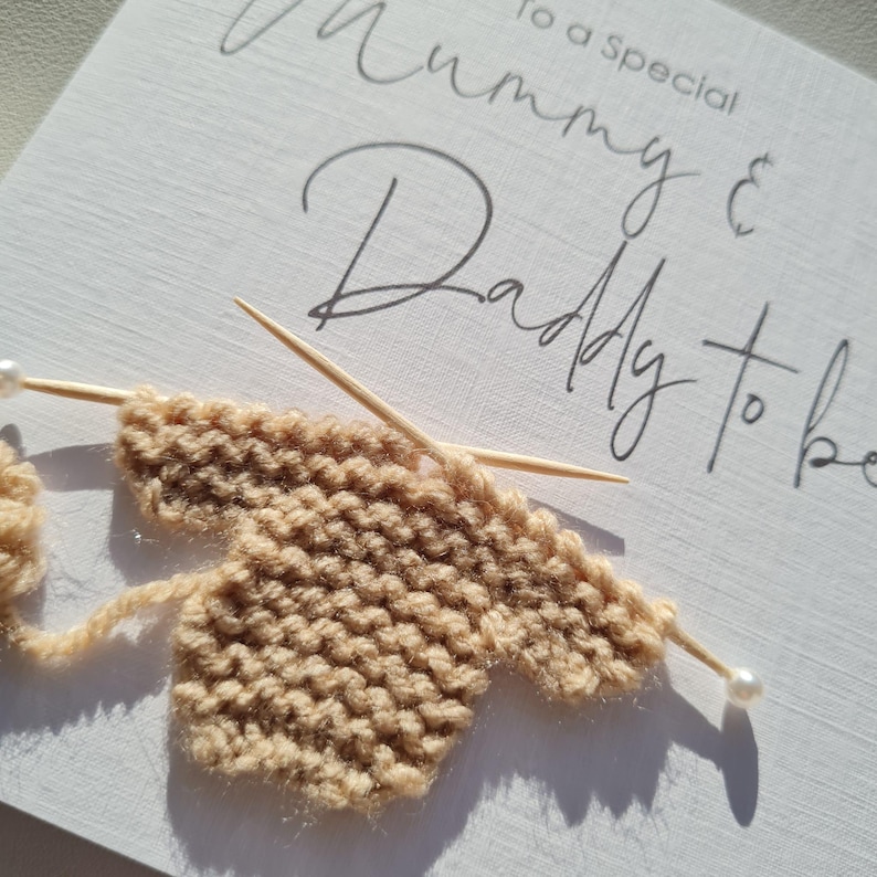 May include: A handmade greeting card with the words "To a Special Mummy & Daddy to be" in elegant script. The card features a small, knitted beige baby garment with knitting needles and pearl-tipped pins, suggesting a baby shower or pregnancy announcement.