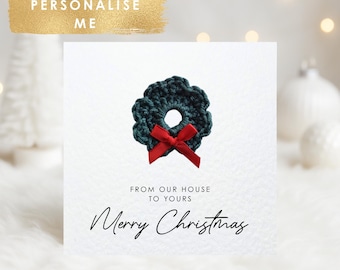 May include: A white Christmas card featuring a dark green crocheted wreath with a red bow. The card includes the text "From our house to yours" and "Merry Christmas". The words "Personalise Me" are in a gold banner.