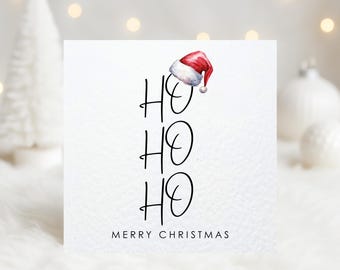 May include: A white Christmas card with the words "HO HO HO" in black script, topped with a watercolor Santa hat. The phrase "MERRY CHRISTMAS" is written at the bottom in a simple font. The background is blurred with soft, warm lighting.