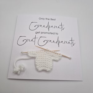 May include: A white card with black text that reads "Only the Best Grandparents get promoted to Great Grandparents". A small white knitted sweater is attached to the card with two wooden skewers.