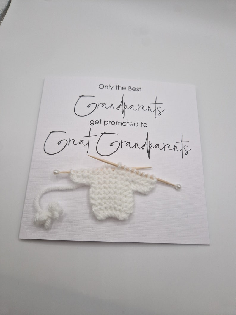 May include: A white card with black text that reads "Only the Best Grandparents get promoted to Great Grandparents". A small white knitted sweater is attached to the card with two wooden skewers.