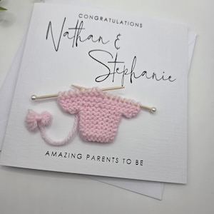 May include: A white card with the words "Congratulations Nathan & Stephanie" in black script. A small pink knitted sweater with knitting needles and the words "Amazing Parents To Be" are also on the card.
