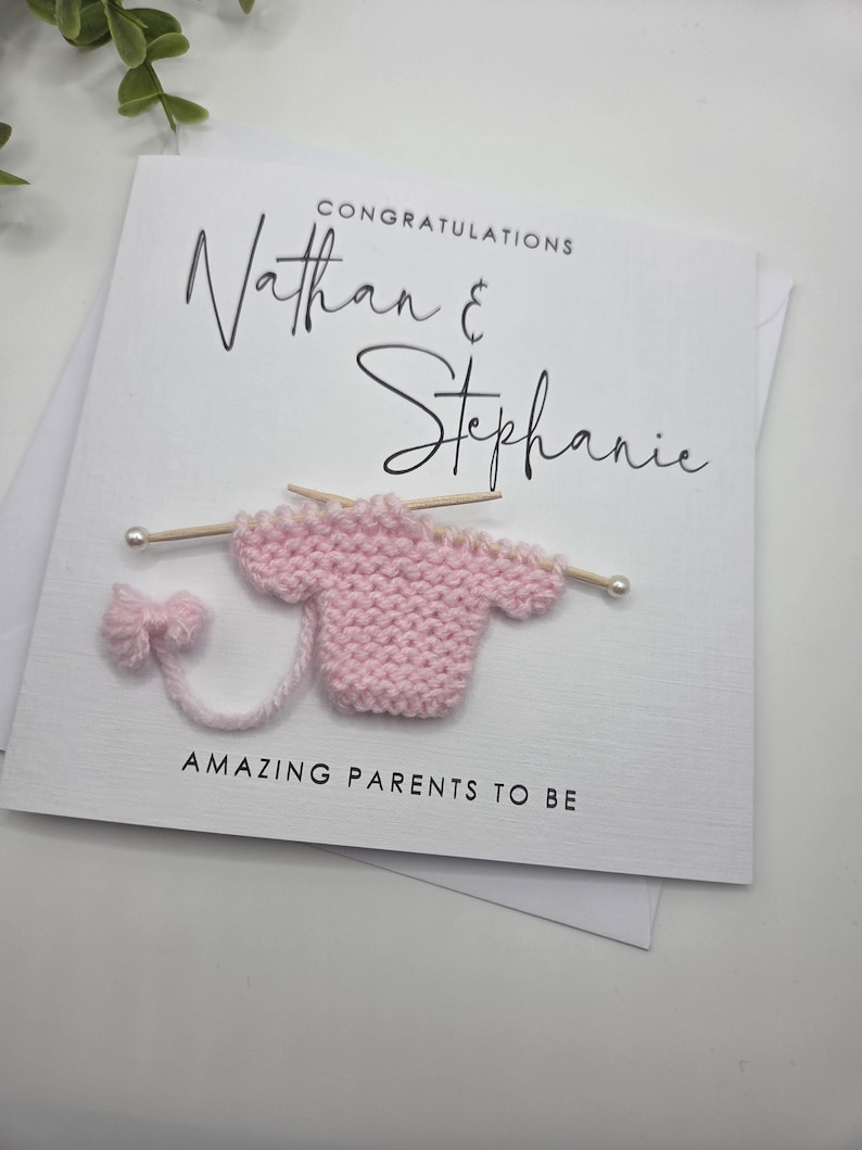 May include: A white card with the words "Congratulations Nathan & Stephanie" in black script. A small pink knitted sweater with knitting needles and the words "Amazing Parents To Be" are also on the card.