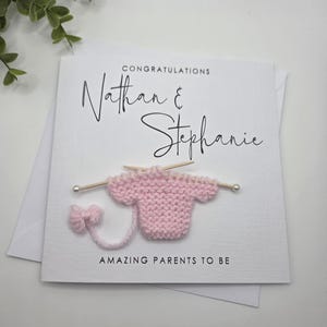 May include: A white card with the names "Nathan & Stephanie" in elegant script, with the word "CONGRATULATIONS" above. A pink knitted baby sweater is attached, with the words "AMAZING PARENTS TO BE" below.