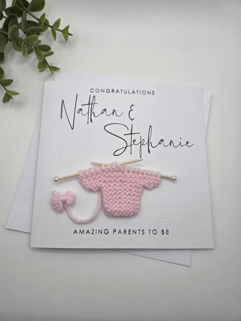 May include: A white card with the names "Nathan & Stephanie" in elegant script, with the word "CONGRATULATIONS" above. A pink knitted baby sweater is attached, with the words "AMAZING PARENTS TO BE" below.