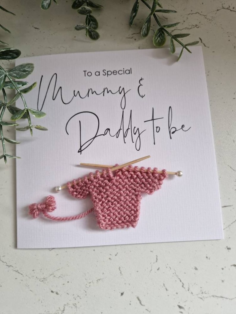 May include: A white card with the text "To a Special Mummy & Daddy to be" in black script. A small pink knitted sweater is attached to the card with wooden knitting needles.