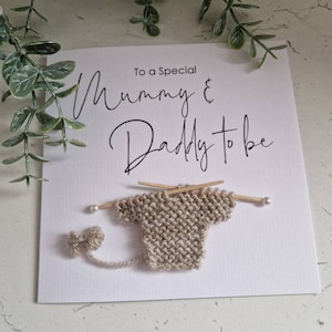 May include: A white card with the text "To a Special Mummy & Daddy to be" in black script. A small knitted baby sweater is attached to the card with two wooden knitting needles.