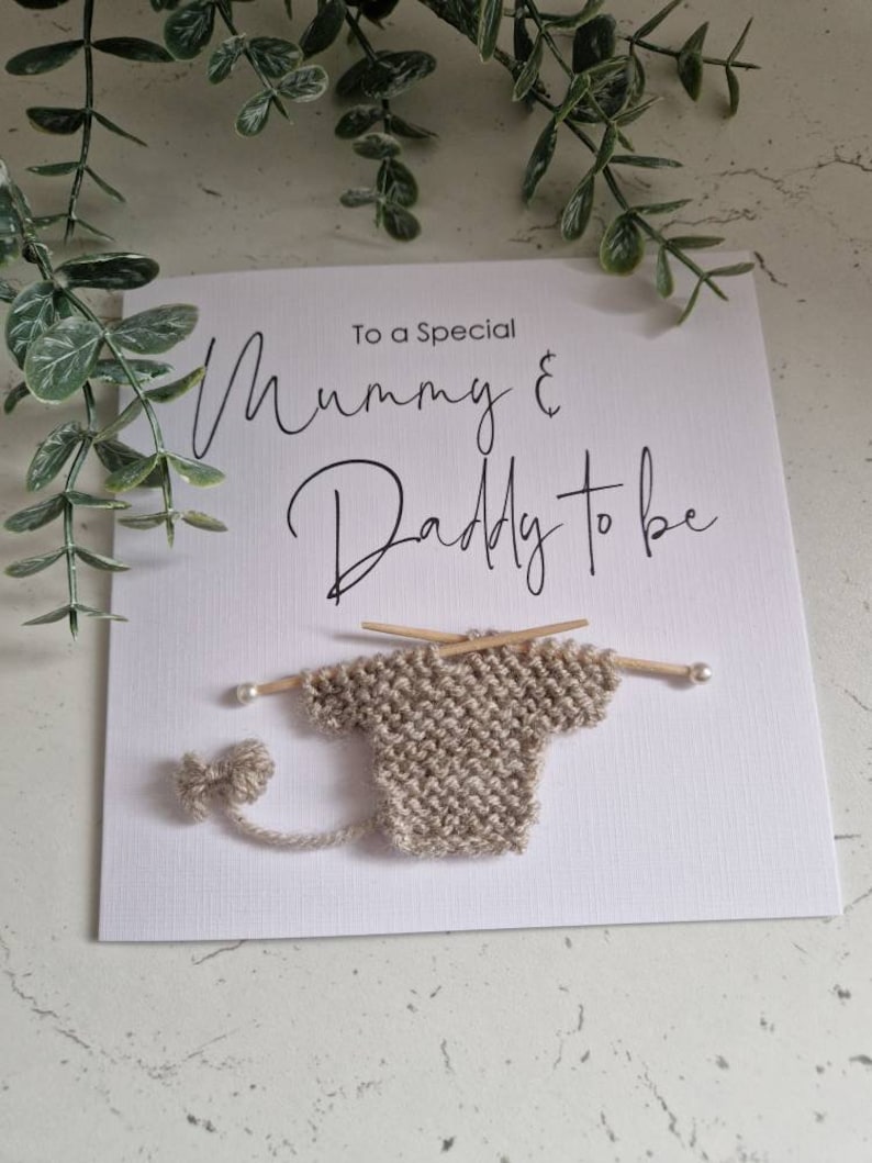 May include: A white card with the text "To a Special Mummy & Daddy to be" in black script. A small knitted baby sweater is attached to the card with two wooden knitting needles.