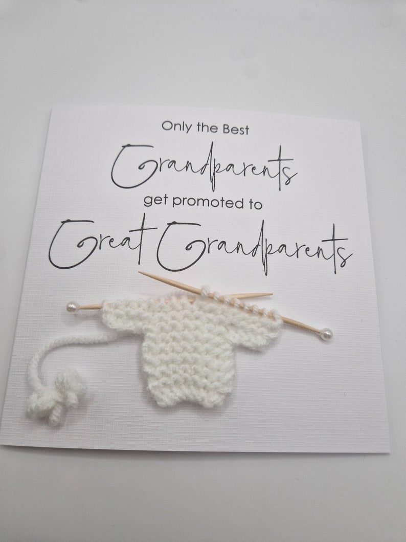 May include: A white card with black text that reads "Only the Best Grandparents get promoted to Great Grandparents". A small white knitted sweater is on the card with two wooden knitting needles and a white yarn ball.