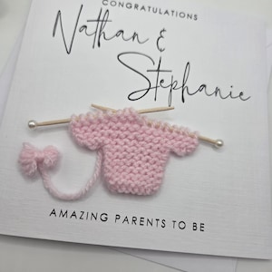 May include: A white greeting card with the names "Nathan & Stephanie" and the word "Congratulations" in black script. A pink knitted baby sweater with knitting needles and a bow are attached to the card. The text "AMAZING PARENTS TO BE" is printed at the bottom.