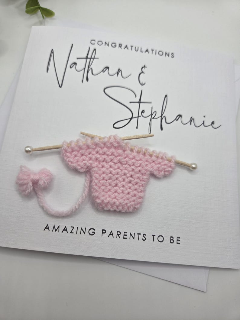 May include: A white greeting card with the names "Nathan & Stephanie" and the word "Congratulations" in black script. A pink knitted baby sweater with knitting needles and a bow are attached to the card. The text "AMAZING PARENTS TO BE" is printed at the bottom.
