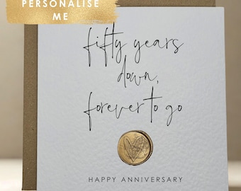 May include: A white anniversary card with the text "fifty years down, forever to go" in black script. A gold-coloured heart-shaped emblem is centered below the text. The words "Happy Anniversary" are printed at the bottom.