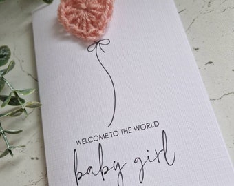 May include: A white card with a pink crocheted heart attached to a string. The card says "Welcome to the world baby girl".