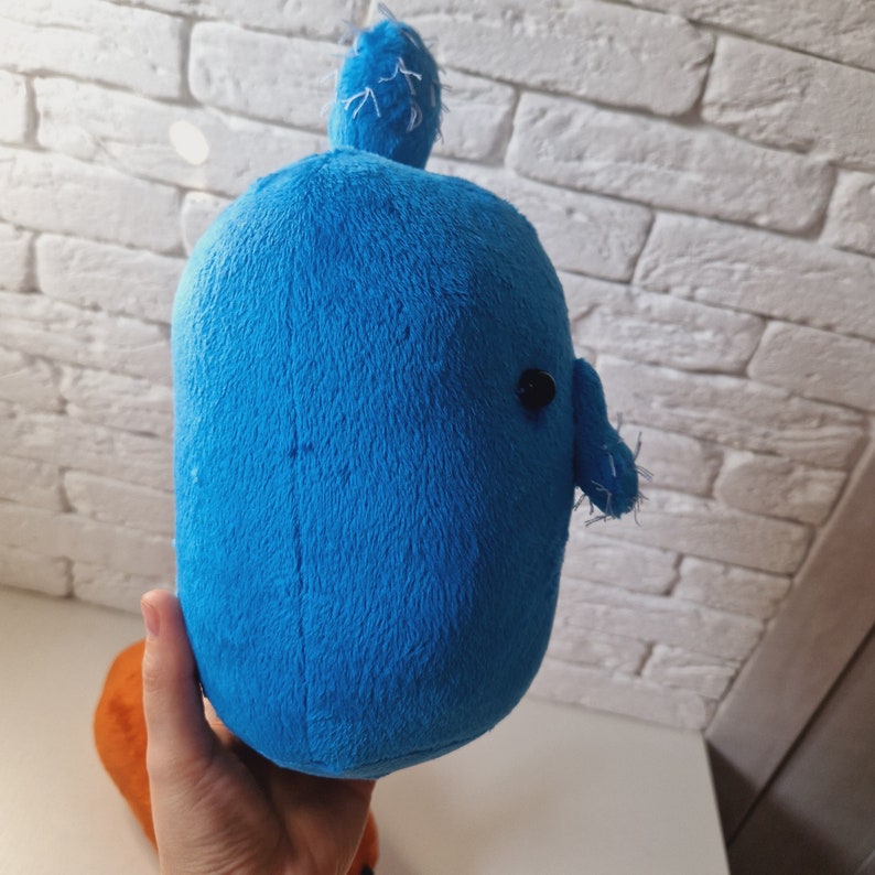 May include: A blue plush toy shaped like a whale with a small black eye. The whale has a textured surface and a small fin on top of its head.