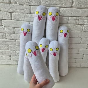 May include: A collection of seven white plush toys, each featuring a cylindrical body, two yellow circular eyes, and a pink V-shaped mouth with small white fangs. The plush toys have embroidered facial details and a soft, fuzzy texture.