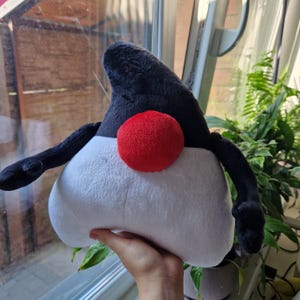 May include: A black and white plush toy shaped like a triangle with a red nose. The toy has two black arms and a black top.