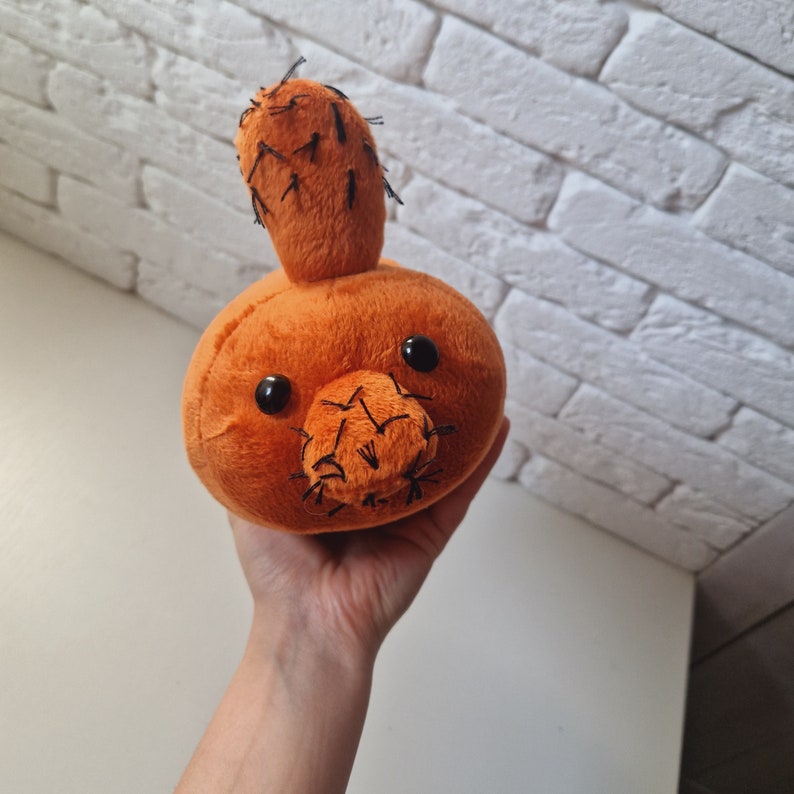 May include: A plush toy shaped like a cactus with a brown body and black stitching for the eyes and mouth. The cactus has a green top with black stitching for the spines.