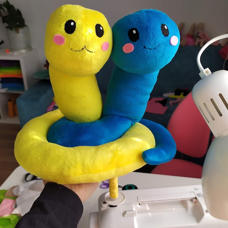 Pu&ograve; includere: Un giocattolo di peluche giallo e blu a forma di due serpenti intrecciati. Il serpente giallo ha un viso sorridente con guance rosa e il serpente blu ha un viso sorridente con guance rosa.