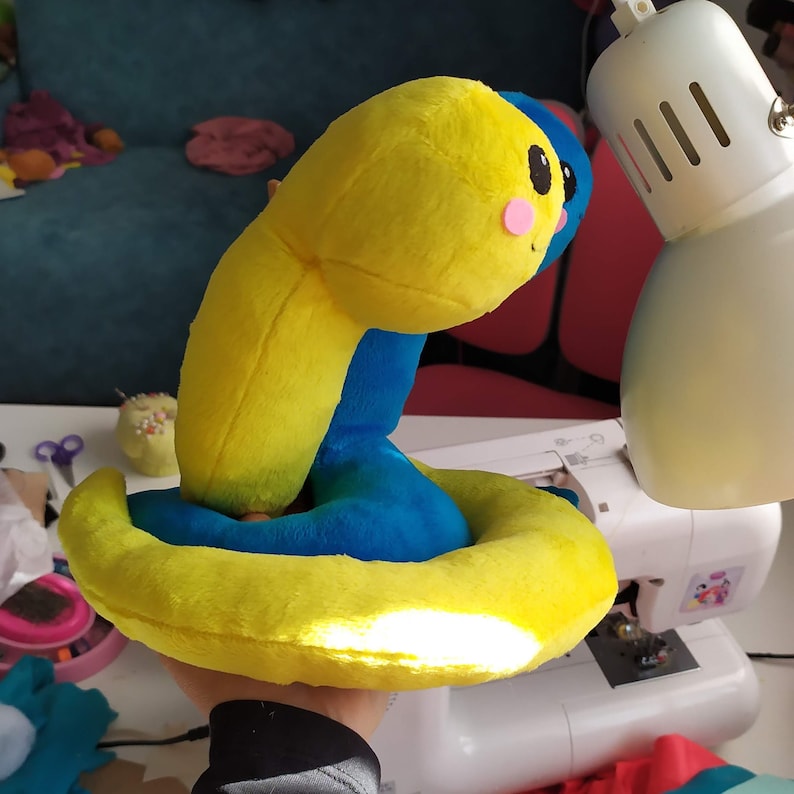 Pu&ograve; includere: Un giocattolo di peluche giallo e blu a forma di serpente. Il serpente &egrave; arrotolato con la coda in bocca. Il giocattolo ha un viso carino con guance rosa e occhi neri.